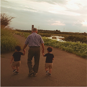 オレンジ色の夕焼け空の中広大な自然を歩いているお爺ちゃんと手をつないでいる二人の子どもの写真